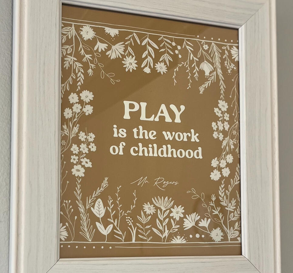 Play is the Work of Childhood "Play is the Work of Childhood" in white font surrounded by white floral elements, in a white frame.