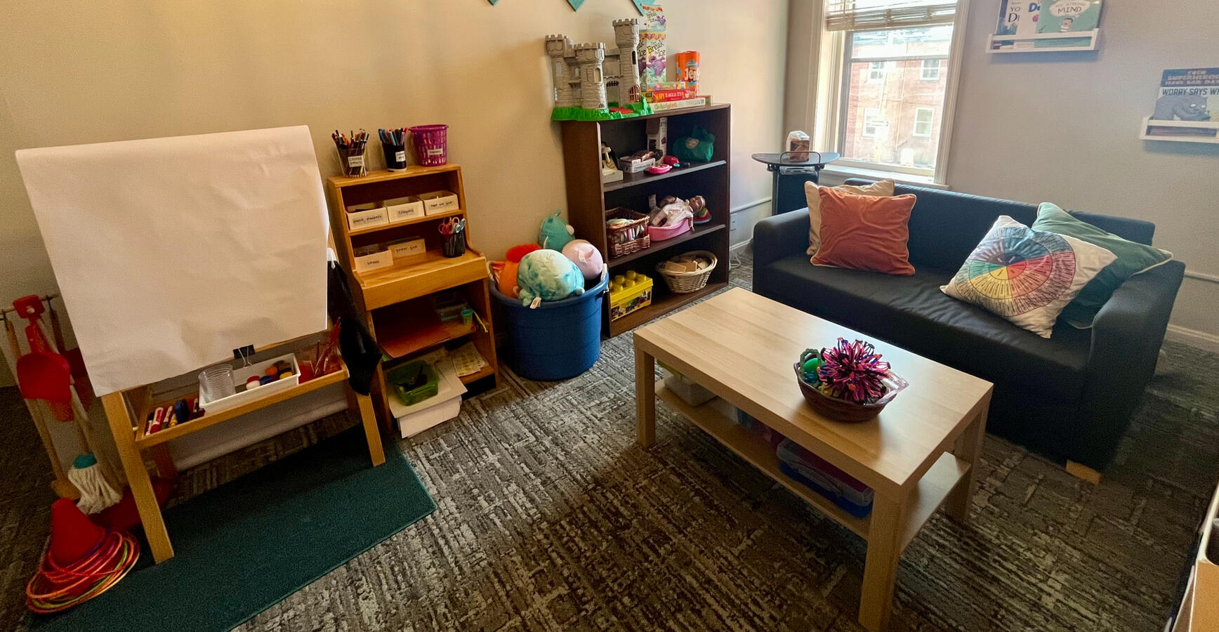 The PAYA Playroom shot at an angle from the wall opposite the entry, including a couch, art easel, and shelves full of toys.