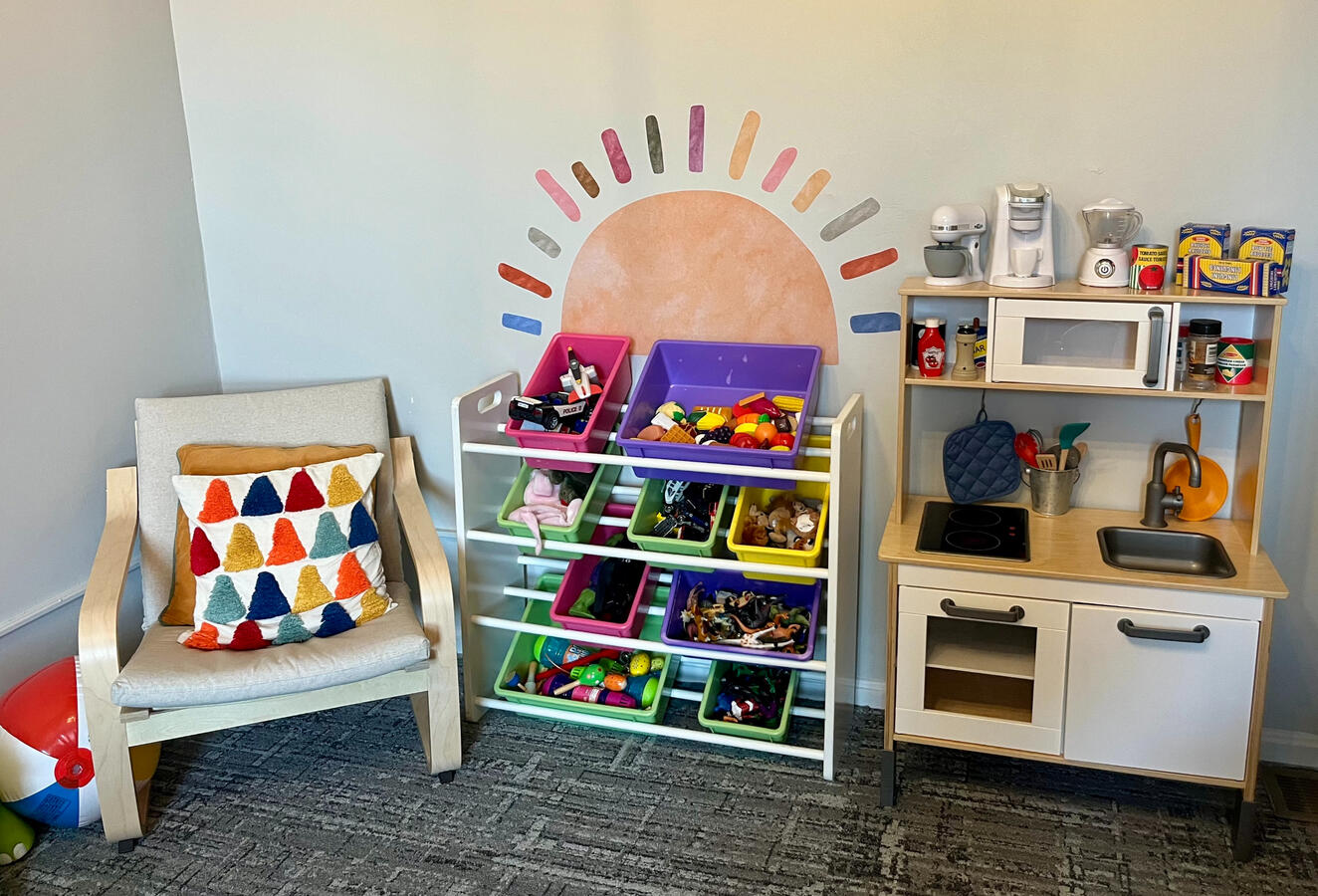 Playroom front wall The front wall of the PAYA Playroom, including a seat, a play kitchenette, a shelf of toys, and a sun mural with colorful rays.